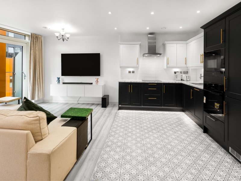 Open Plan Living Area Featuring A Neutral Sofa, Wall Mounted TV, And A View Into A Modern Black And White Kitchen.